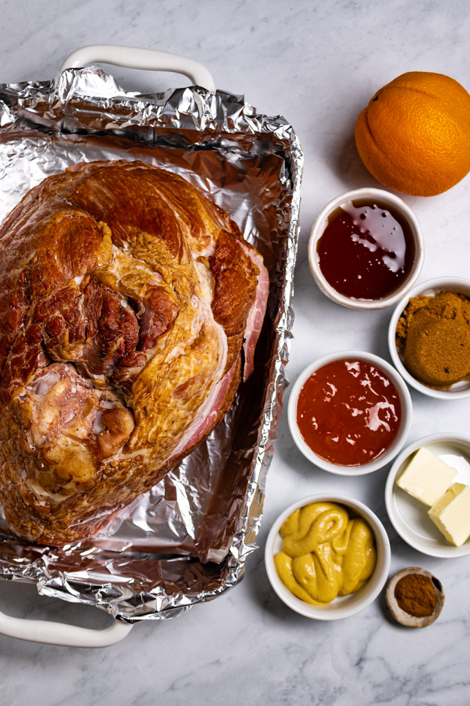 Uncooked spiral ham in a baking dish lined with foil. Ingredients in small bowls off to the right side: Dijon mustard, apricot jam, honey, brown mustard, and an orange.