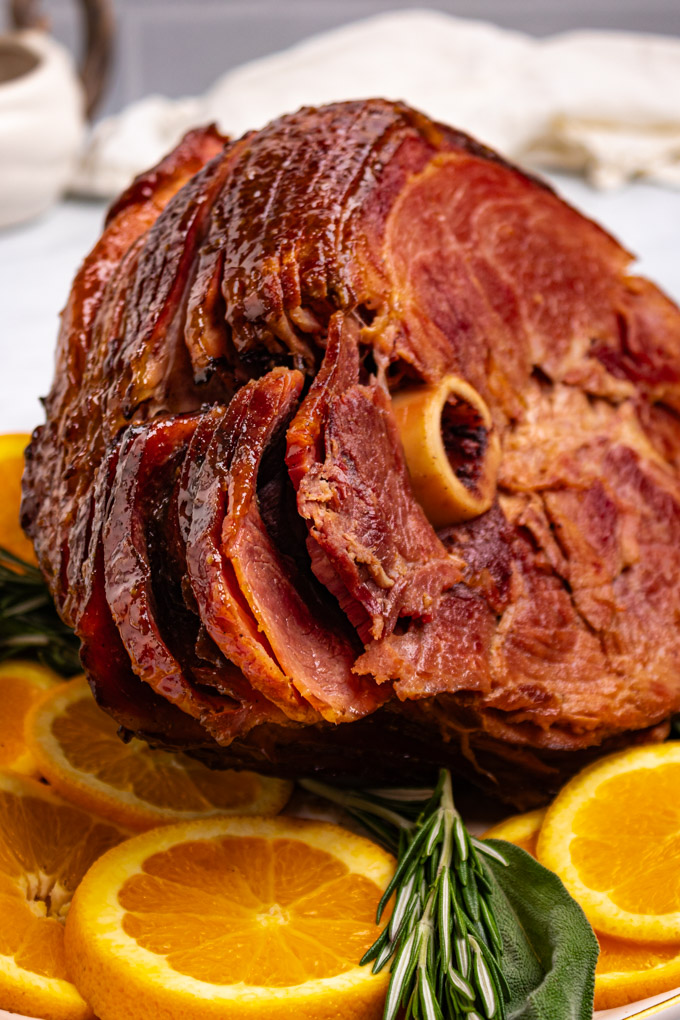 Closeup angled photo of honey glazed ham. Orange slices, and sprigs of rosemary, and sage are garnishing the platter.