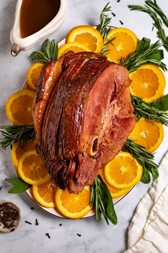 Overhead photo of honey glazed ham on a platter. The platter is garnished with slices of orange, rosemary, and sage sprigs.