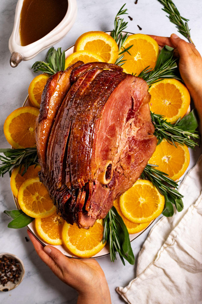 Overhead photo of honey glazed ham on a platter with orange slices, sage, and rosemary garnishing the platter. Hands are holding the platter. A gravy boat with pan drippings is in the upper left corner of the photo..