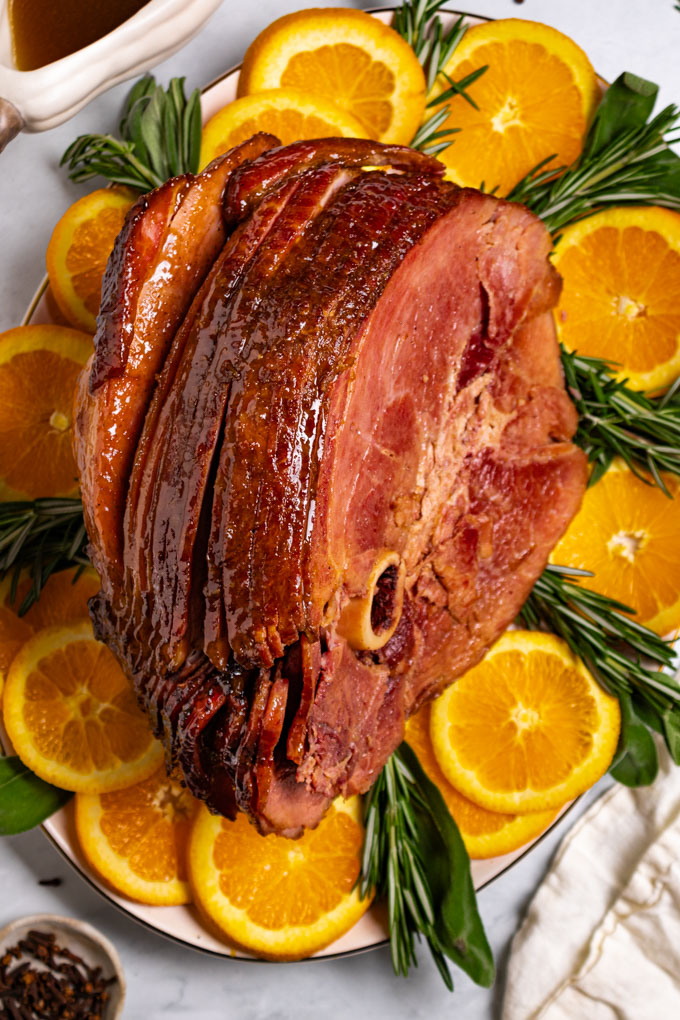 Overshot photo of honey glazed ham on a platter with oranges, rosemary, and sage garnishing the platter. Cloves are in a small bowl in the bottom left corner.