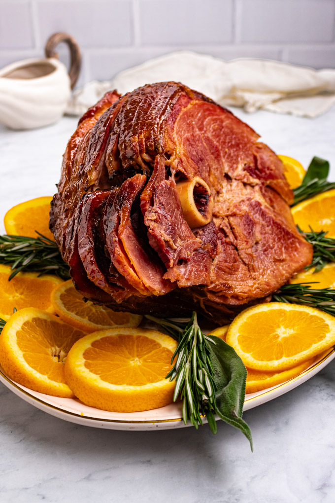 Angled shot of honey glazed ham on a serving platter. Sliced oranges, and sprigs of rosemary, and sage are garnishing the ham on the platter.