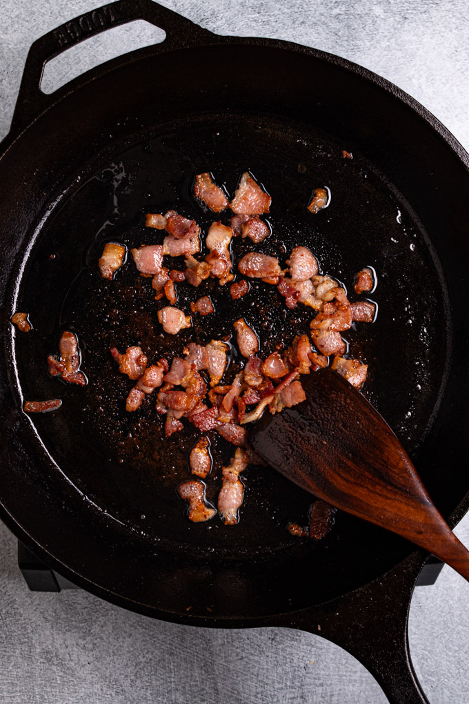 Overhead photo of chopped bacon cooked in a skillet. A wooden spatula is stirring the bacon.