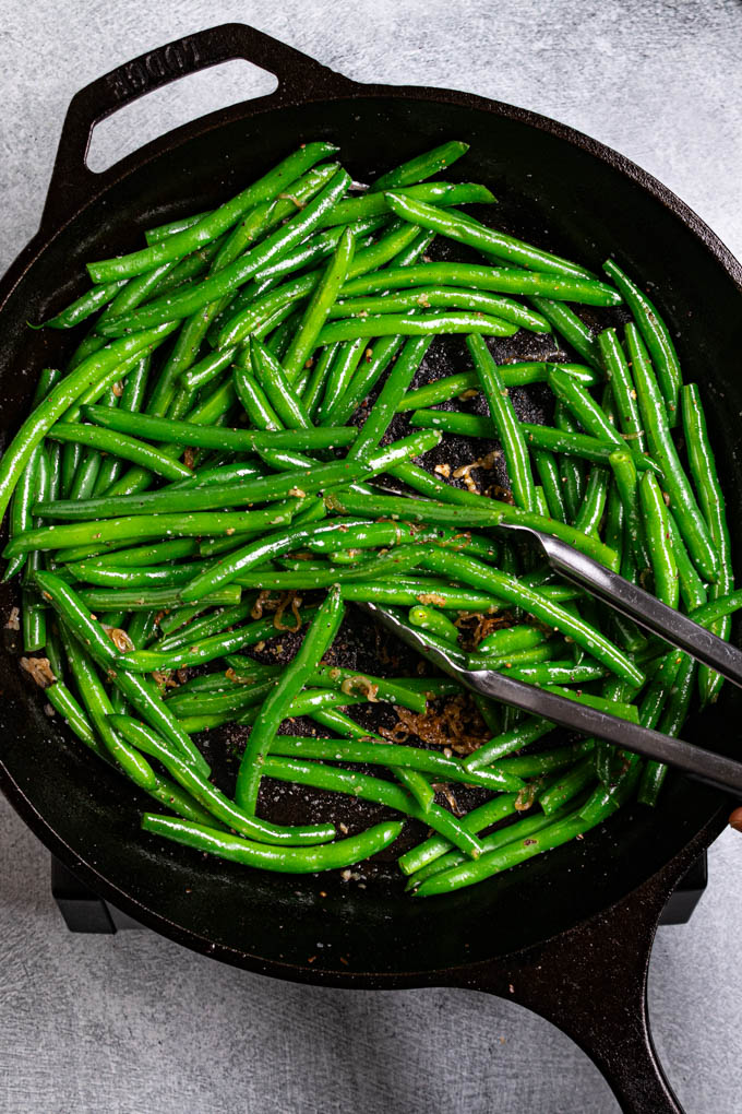 Overhead photo of green beans in a cast iron skillet. Tongs are tossing the green beans in the pan.