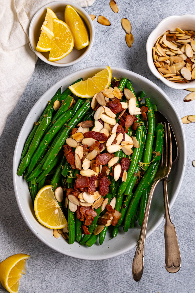 Overhead shot of green beans almondine with bacon. Green beans are in a serving bowl with bacon, and almonds sprinkled on top. There two lemon wedges on the left side, along with a serving fork and spoon to the right of the bowl. A small bowl of lemon wedges are in the upper left corner, sliced almonds are in the upper right corner of the photo too.