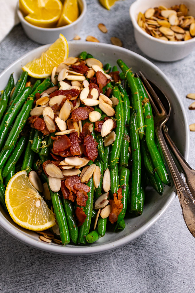 Angled, close up photo of green beans almondine with bacon. Green beans are in a serving bowl with bacon, and almonds sprinkled on top. There two lemon wedges on the left side, along with a serving fork and spoon to the right of the bowl. A small bowl of lemon wedges are in the upper left corner, sliced almonds are in the upper right corner of the photo too.
