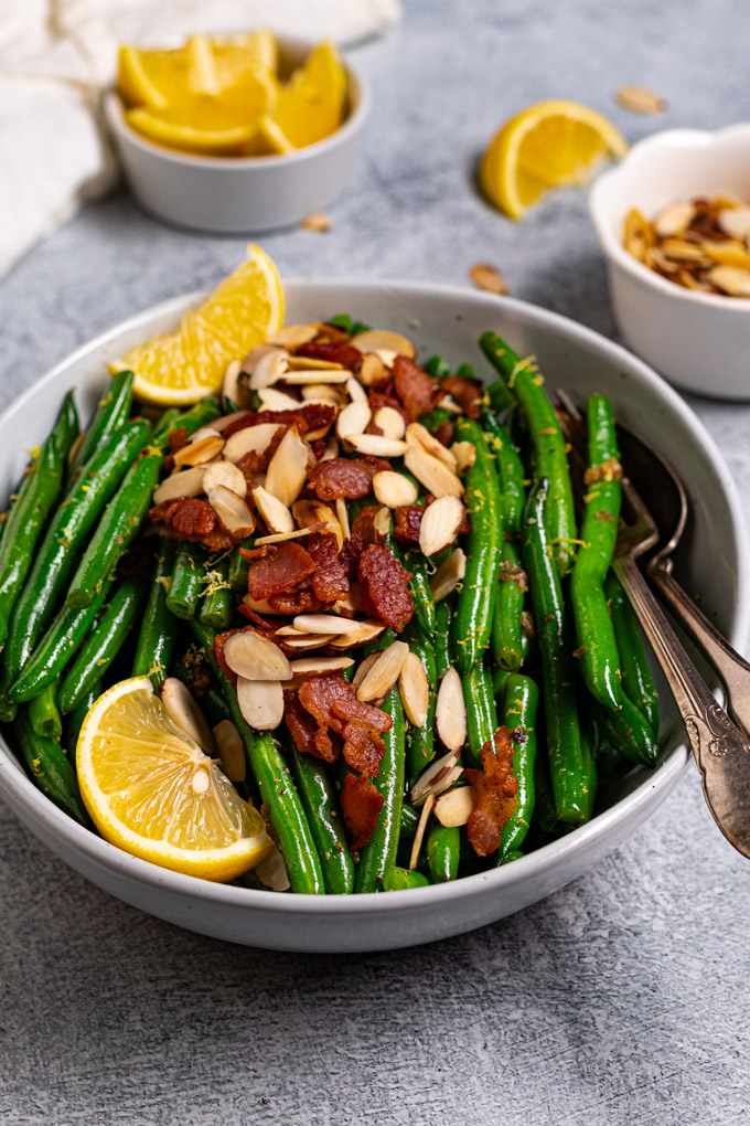 Angled photo of green beans almondine with bacon. Green beans are in a serving bowl with bacon, and almonds sprinkled on top. There two lemon wedges on the left side, along with a serving fork and spoon to the right of the bowl. A small bowl of lemon wedges are in the upper left corner, sliced almonds are in the upper right corner of the photo too.