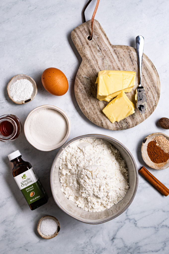 Overhead photo of ingredients. Flour in a bowl, sugar in a bowl, a bottle of almond extract, cinnamon, and nutmeg, an egg, and butter is on a small marble platter with a knife.
