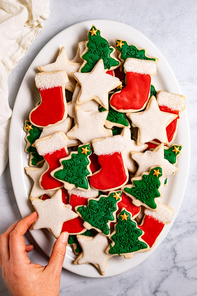 Overhead photo gluten free sugar cookie cut outs layered on an oval platter, on a gray marble background. The cookies are decorated for Christmas, they are cut into stars, stockings, and Christmas trees. They are decorated in red, white, and green icing. There is one hand in the bottom left corner holding a star cookie that is decorated with white icing.
