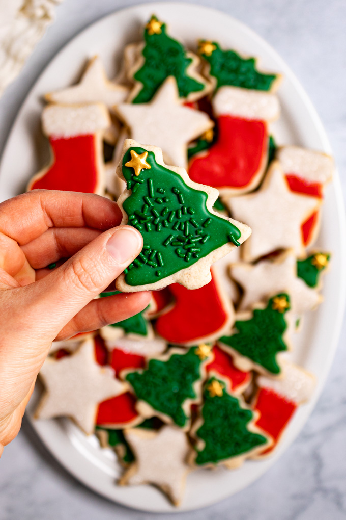 Overhead photo gluten free sugar cookie cut outs layered on an oval platter. The cookies are decorated for Christmas, they are cut into stars, stockings, and Christmas trees. They are decorated in red, white, and green colors. A hand is holding a Christmas tree cookie up close to the camera, which is in focus. The cookies on the platter are out of focus to create depth of field. There are small bowls off to the side that have various sprinkles in them.