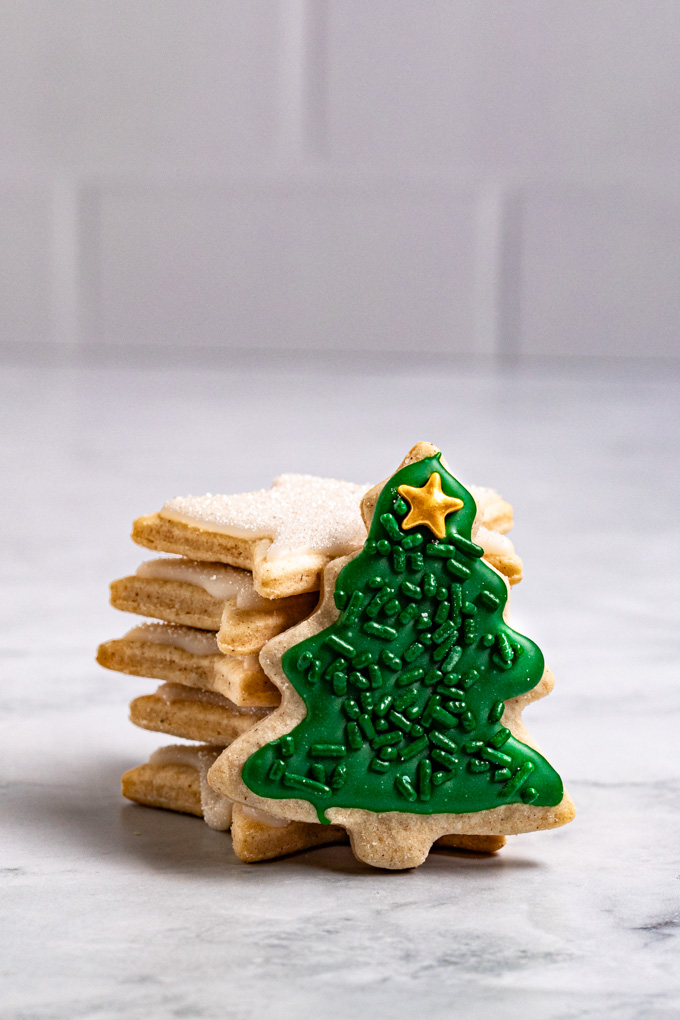 A close up angled photo of a gluten free sugar cookie cut out in the shape of a Christmas tree with green icing on it, green sprinkles, and a gold star sprinkle on the top. The Christmas tree cookie is standing upright and is propped up by a stack of star cookies that are decorated in white icing. There is a simple gray marble background and a gray subway tile backdrop behind the cookies.