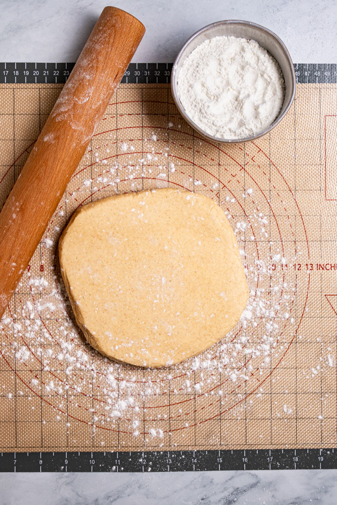 Overhead photo of a disc of gluten free sugar cookie dough on a floured pastry mat that is on a marble surface. A rolling pin is off to the left side, and a small bowl of dusting flour is to the upper right side.