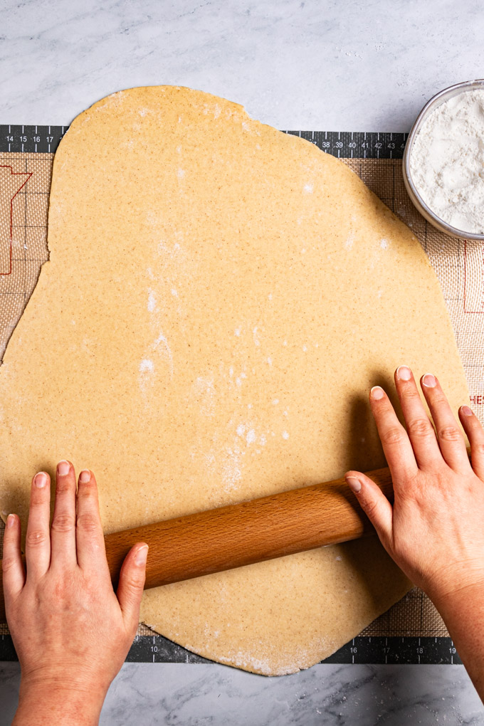 Overhead photo of gluten free cookie dough being rolled out with a roiling pin. Two hands are rolling the rolling pin. A small bowl of dusting flour is in the upper right hand corner.