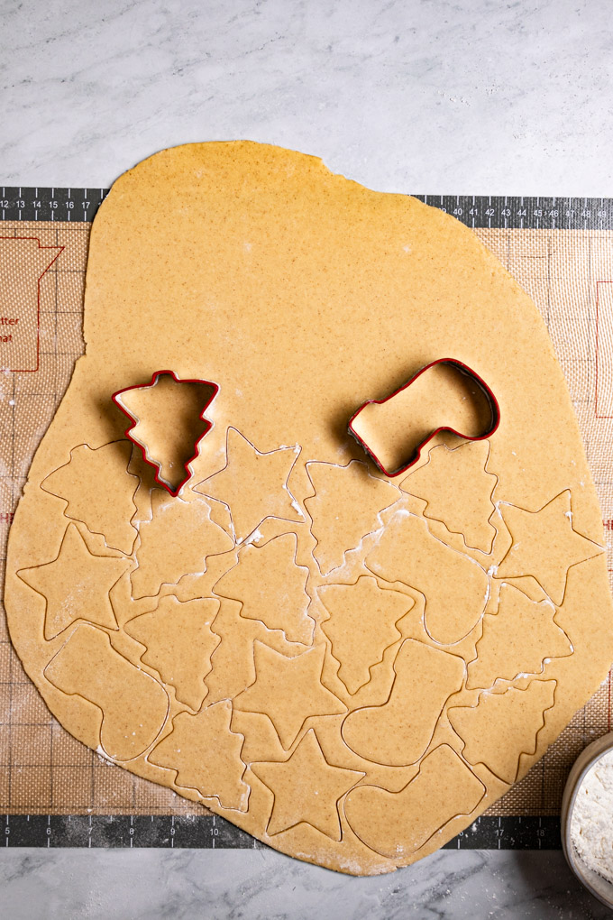Overhead shot of gluten free sugar cookie dough that has been rolled out. There are outlines of cookie shapes in stockings, stars, and Christmas trees. A Christmas tree cookie cutter, and a stocking cookie cutter are laying on the surface of the cookie dough.