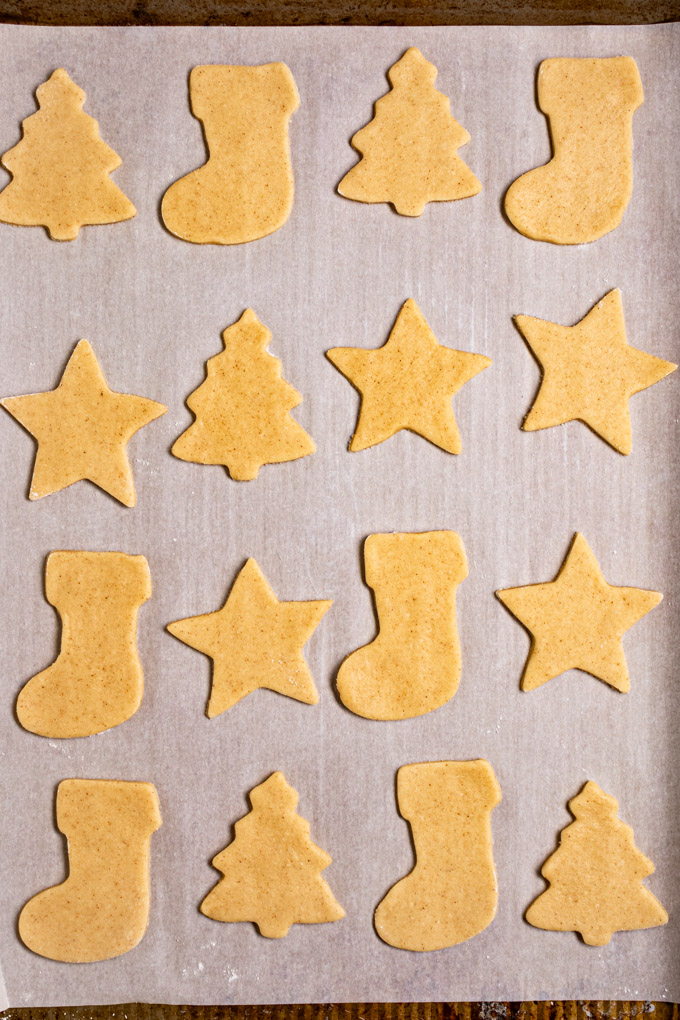 Closeup overhead photo of unbaked sugar cookie cut outs on a baking pan that's lined with parchment paper. The shapes of the cookies are cut out into stars, Christmas trees, and stockings.