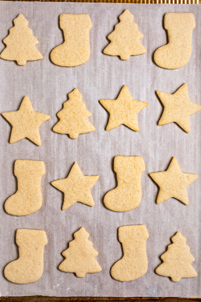 Closeup overhead photo of baked sugar cookie cut outs on a baking pan that's lined with parchment paper. The shapes of the cookies are cut out into stars, Christmas trees, and stockings.