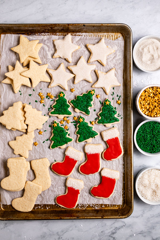 Overhead photo of gluten free sugar cookies lined up on a baking pan with parchment paper. The sugar cookies are cut into stars, Christmas trees, and stockings. Some of the cookies are un-decorated, and some are decorated with white, green, and red icing colors with sprinkles, and sanding sugar for decoration. Coordinated sprinkles are in small bowls off to the right of the cookie sheet.
