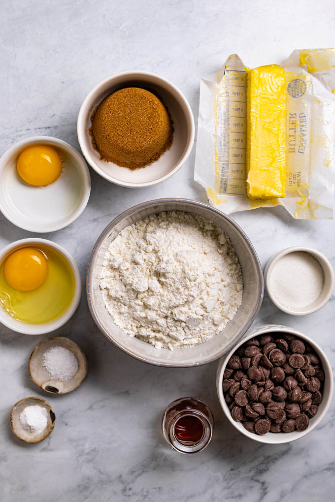 Ingredients in bowls: flour, chocolate chips, sugar, brown sugar, butter, a whole egg in a bowl, an egg yolk in a bowl, salt, baking powder, and vanilla.