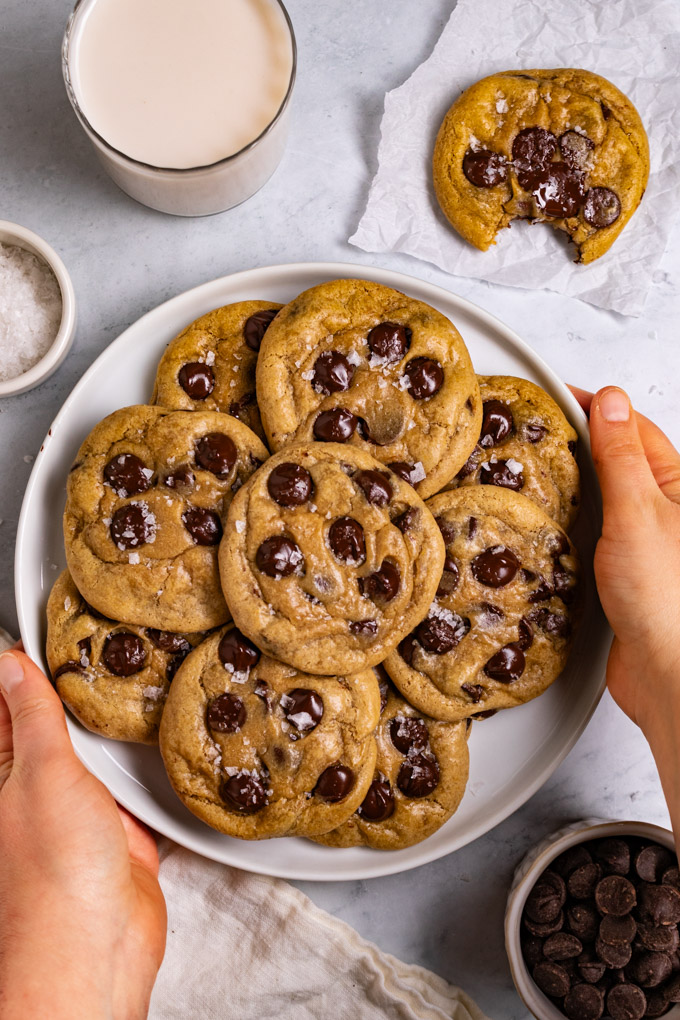 A plate full of gluten free chocolate chip cookies on a light marble background. Two hands are holding the plate of cookies. There is a cookie in the upper right hand corner with a bite taken out of it. There is also a glass of milk in the upper left corner, along with a small bowl of chocolate chips in the lower right corner.