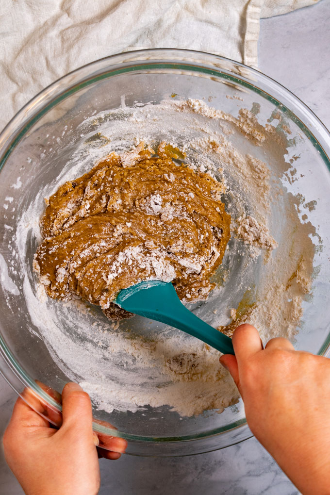 Cookie batter is being mixed in a mixing bowl. A hand is holding the spatula, while another hand is holding the glass mixing bowl.