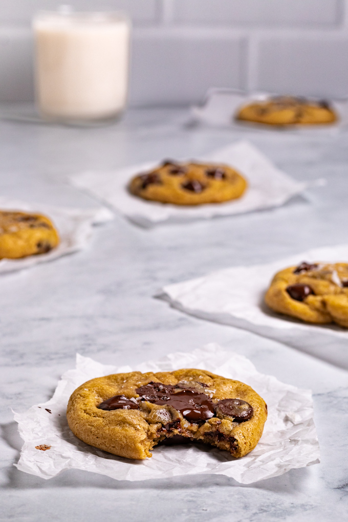 Angled shot of gluten free chocolate chip cookies. One cookie is in the center with a bite taken out of it. There are 4 more cookies scattered behind the front cookie. A glass of milk is in the background as well. 