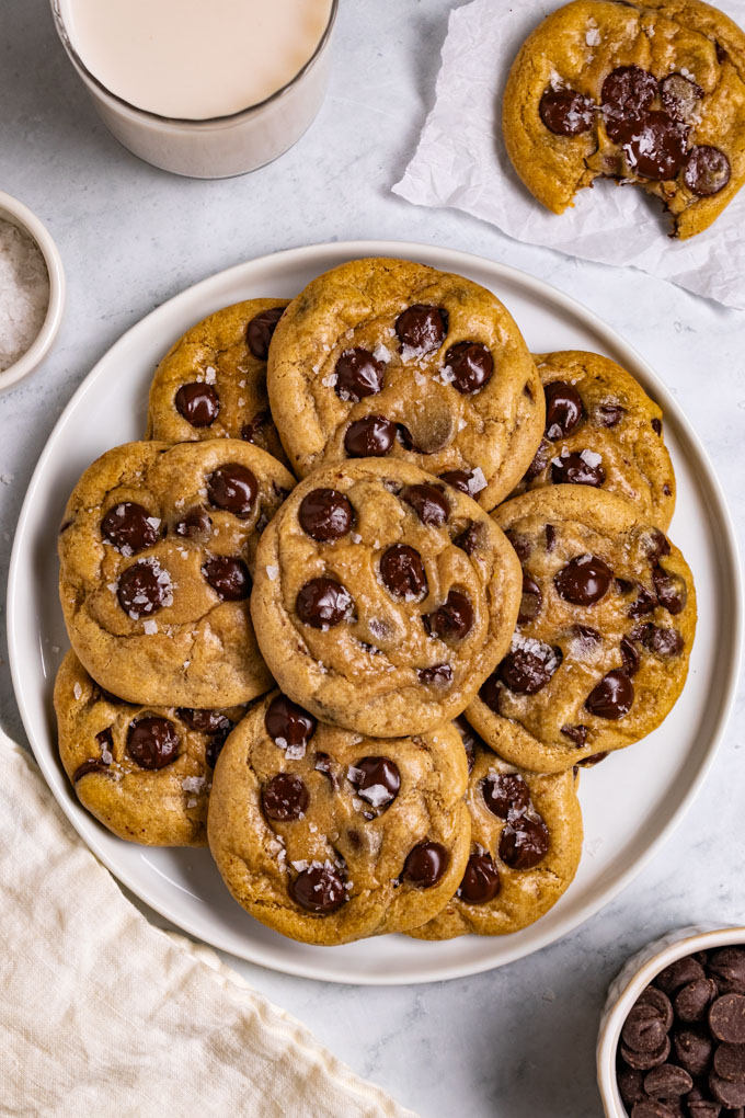 A plate full of gluten free chocolate chip cookies on a light marble background. There is a cookie in the upper right hand corner with a bite taken out of it. There is also a glass of milk in the upper left corner, along with a small bowl of chocolate chips in the lower right corner.