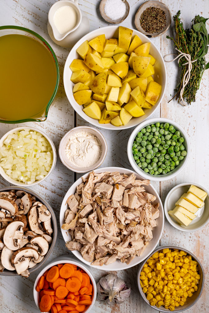 Overhead photo of ingredients in bowls - cut up chicken, frozen peas, cut up potatoes, corn, cut up carrots and mushrooms, diced onion, chicken broth, and an herb bundle. This scene is photographed on a white distressed wood surface. #foodphotogrpahy