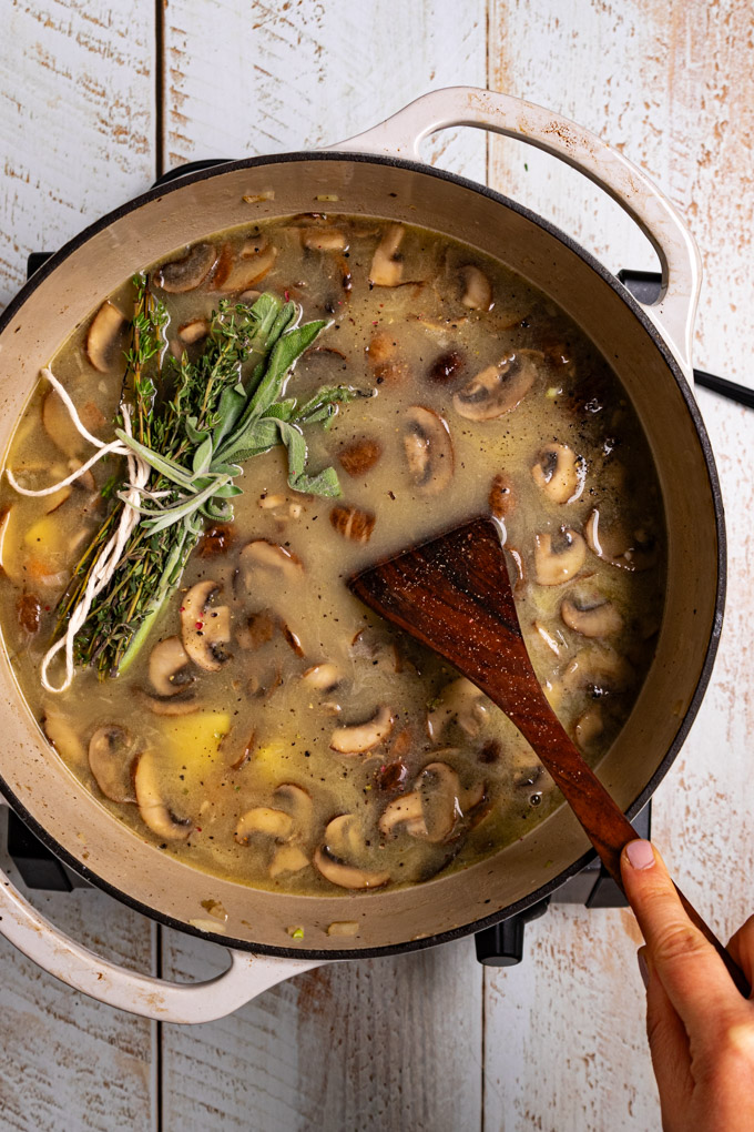 Overhead photo of a white Dutch oven with soup in it. There are lots of mushrooms floating on the surface with a bundle of herbs. A hand is holding a wooden spoon in the soup. The scene has been photographed on a distressed white wood plank surface. #foodphotography