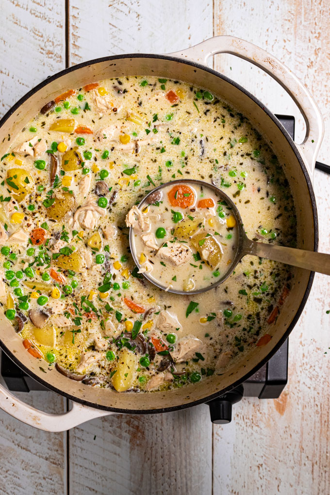 An overhead phot of a white Dutch oven with chicken pot pie soup in it. The soup is creamy with chunks of potatoes, carrots, peas, and mushrooms. It is garnished with fresh parsley and black pepper. A ladle is spooning some of the soup up to the camera. The scene has been photographed on a distressed white wood plank surface. #foodphotography