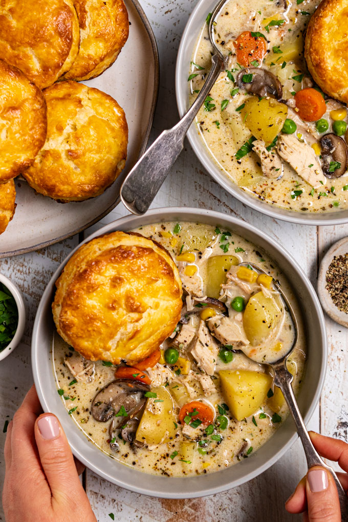 Overhead photo of chicken pot pie soup with biscuits in a gray bowl. A biscuit is resting in the soup. The soup is creamy, there are chunks of potato, carrots, mushrooms, chicken, and peas in the soup. A hand is touching the left side of the bowl, along with another hand holding a spoon in the soup. Another bowl of soup is in the upper right hand corner, and a plate of biscuits is in the upper left corner. The scene has been photographed on a distressed white wood plank surface. #foodphotography