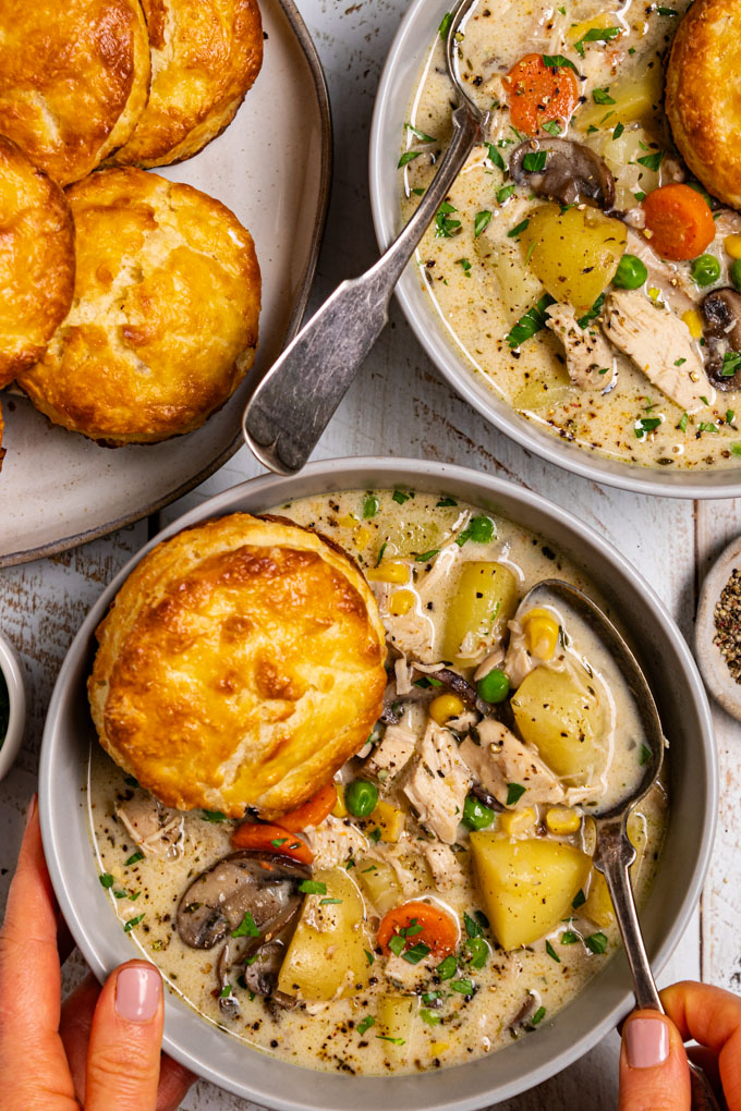 An overhead, close up photo of chicken pot pie soup with biscuits in a gray bowl. A biscuit is resting in the soup. The soup is creamy, there are chunks of potato, carrots, mushrooms, chicken, and peas in the soup. A hand is touching the left side of the bowl, along with another hand holding a spoon in the soup. Another bowl of soup is in the upper right hand corner, and a plate of biscuits is in the upper left corner. The scene has been photographed on a distressed white wood plank surface. #foodphotography