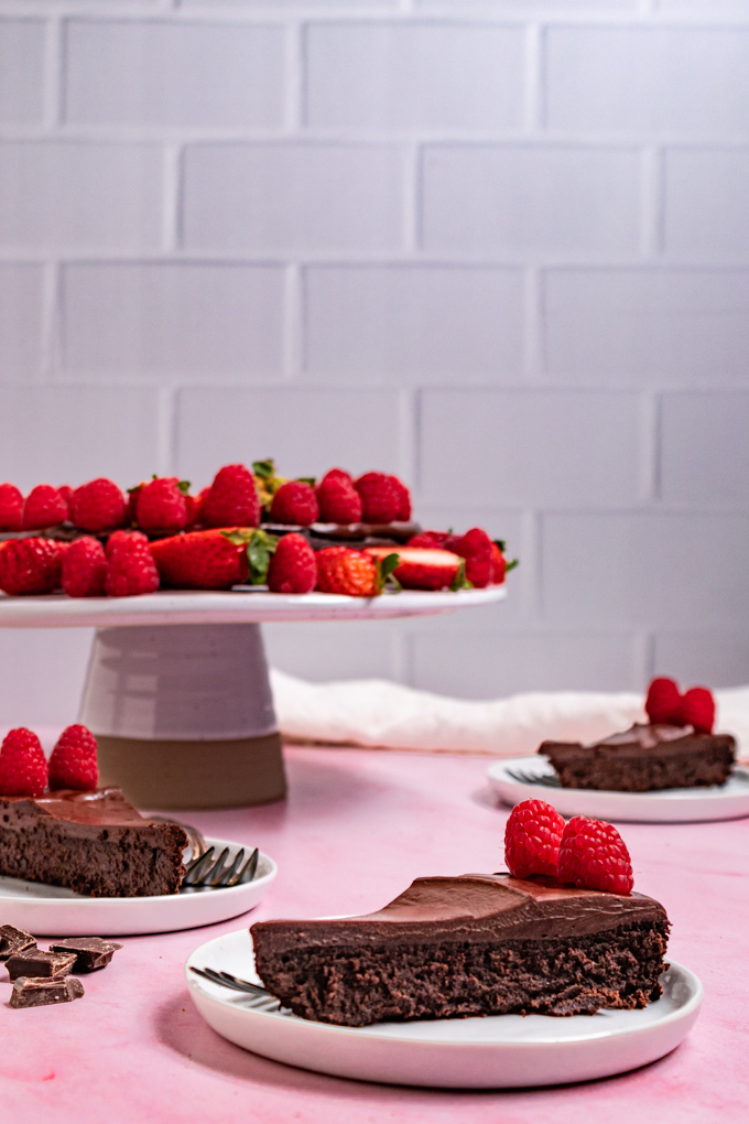 Straight on photo of flourless chocolate cake with ganache. The cake is on a cake stand in the background decorated with fresh raspberries and strawberries. A slice of the cake is in the foreground on a small plate, along with 2 more slices of cake in the background, one to the left, and to the right. #foodphotography