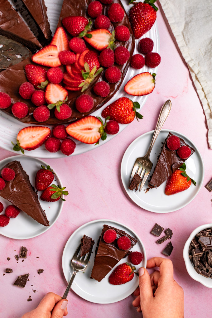 Overhead photo of flourless chocolate cake with ganache - the cake is in the upper left corner on a cake stand, decorated with fresh raspberries and strawberries. There are 3 slices of the cake on small plates in the foreground. Some of the slices have bites taken out of the cake. One of the plates has a hand holding the plate, along with the fork that has a bite of cake on it. #foodphotography
