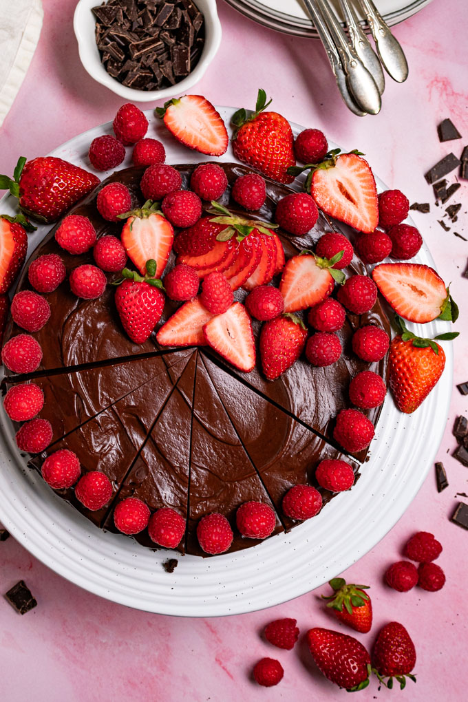 Overhead photo of flourless chocolate cake with ganache on a white cake stand, with a pink marble background. The cake is decorated with fresh raspberries and strawberries. Part of the cake is sliced into sliced. #foodphotography