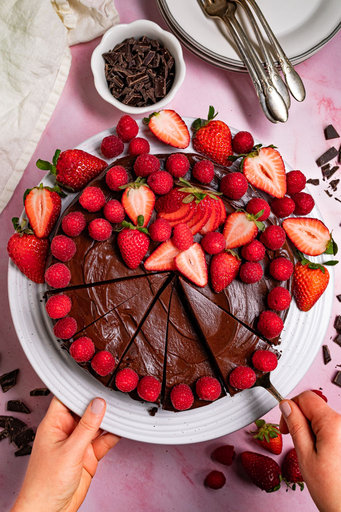 Overhead photo of flourless chocolate cake with ganache on a white cake stand, with a pink marble background. The cake is decorated with fresh raspberries and strawberries. Part of the cake is sliced into, a hand is holding the cake stand, and another hand is holding a cake server that is lifting up one slice of the cake. #foodphotography