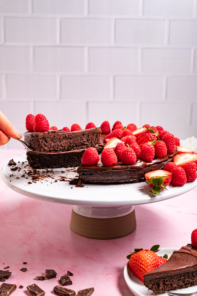 Angled photo of flourless chocolate cake with ganache. The cake is on a cake stand, part of it is sliced up and a single slice of the cake on a cake server with, in the air. A small plate with a slice of cake on it is in the front of the cake stand. Fresh raspberries and strawberries are decorating the cake. #foodphotography