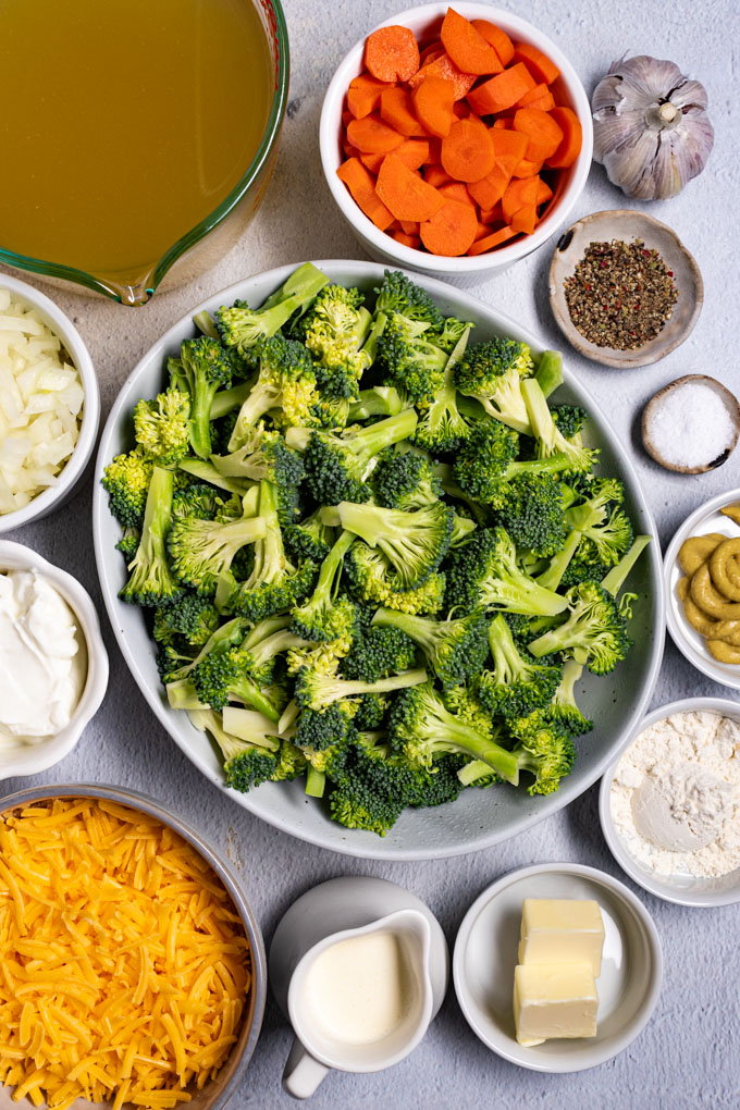 Overhead photo of ingredients in bowls: broccoli florets, shredded cheddar cheese, chopped onion, sliced carrots, garlic cloves, chicken broth, cream, butter, and flour. 