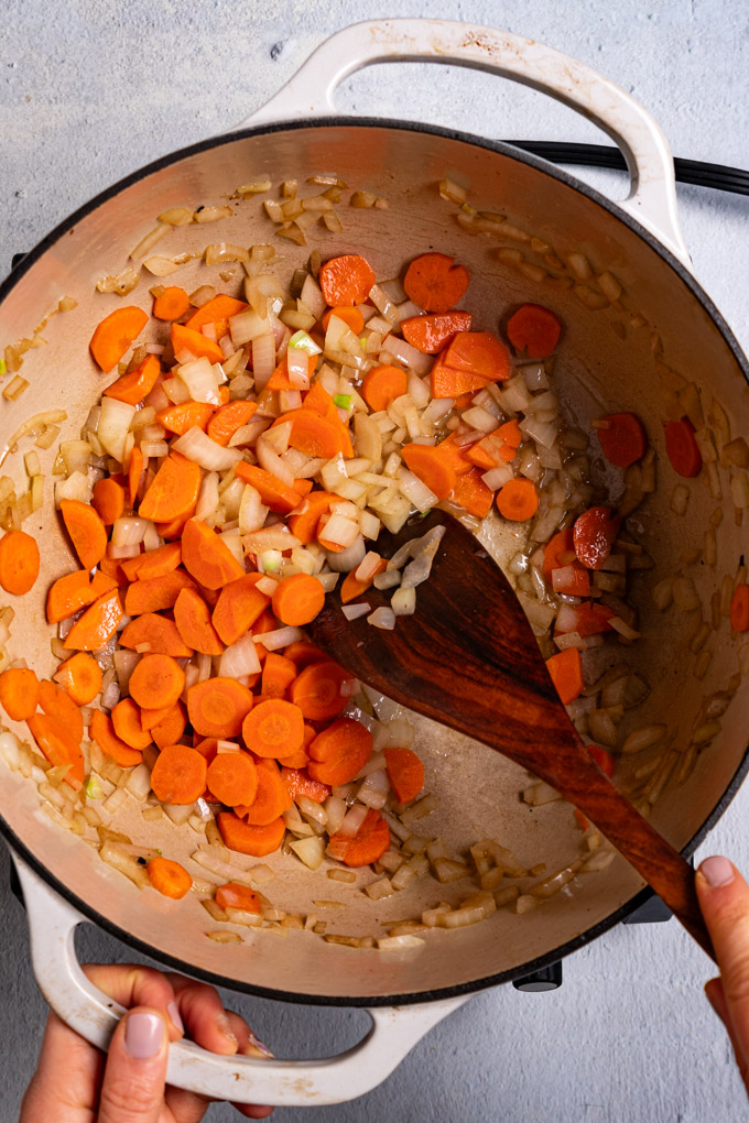 Overhead shot of carrots and onions being sautéed in a white Dutch oven. A hand is holding one of the handles of the Dutch oven while another hand is holding a wooden spoon and is stirring the ingredients in the pot. #foodphotography