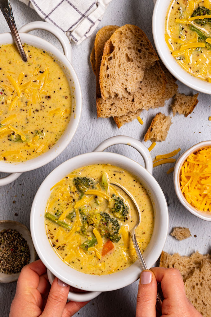 Overhead photo of healthy broccoli cheddar soup in a white bowl. A hand is holding a spoonful of the soup. There is a bowl of soup in the upper left corner as well, and another bowl of soup in the upper right corner. Slices of bread are scattered through the scene. #foodphtoography