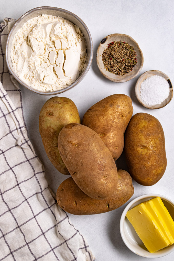 Overhead photo of ingredients on a subtle gray background. Russet potatoes are piled in the center of the photo. A bowl of flour is in the upper left, black pepper, and salt to the right of the flour, and slices of butter in the lower right of the photo. To the left is a white and blue checkered napkin. #foodphotography