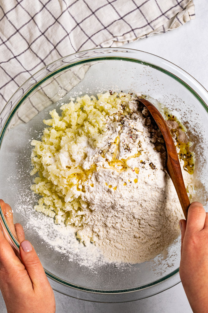 Overhead photo of a glass mixing bowl with ingredients in it. Mashed up potato, flour, melted butter, and salt and pepper. A hand is holding the left side of the bowl, and another is holding a wooden spoon in the mixture, on the right side. 