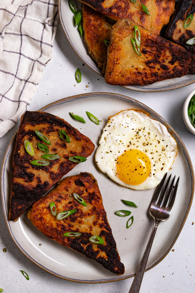 Overhead photo of two Irish potato farls on shot on gray plate with with an over easy egg next to the farls. There are green onions sprinkled on top of the farls. Another plate of farls stacked up are in the upper right corner of the photo. There is a white, and blue checkered striped napkin in the upper left corner. The entire scene is shot on a subtle gray background. #foodphotography